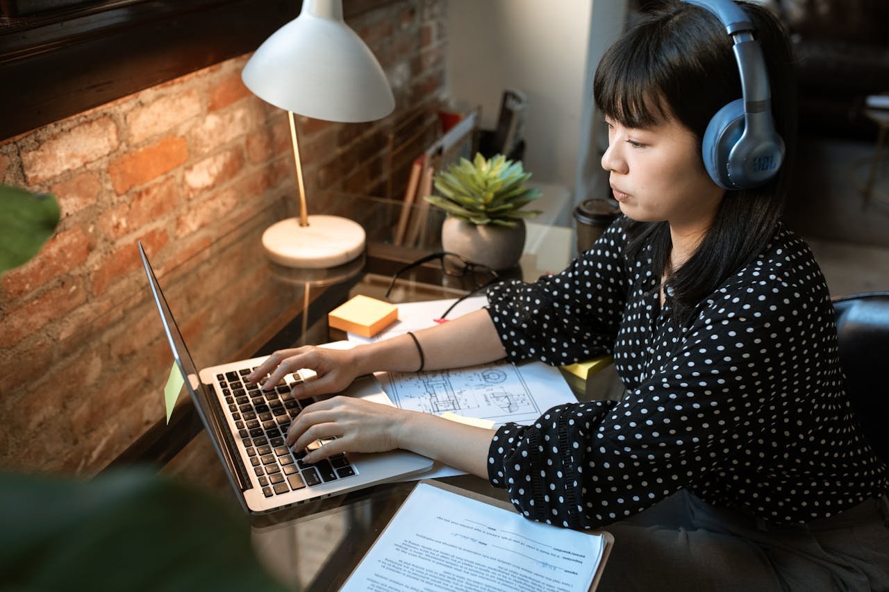 why-choose-us Focused woman wearing headphones, working on a laptop in a cozy home office setup with natural light.