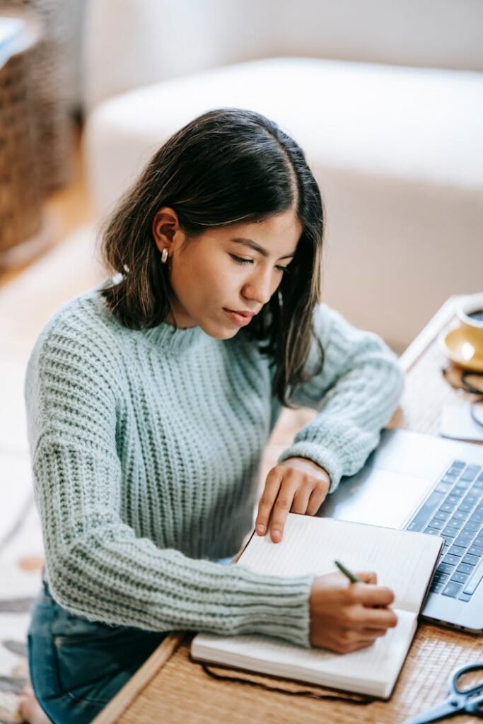 pexels-photo-6347908 Young Hispanic female student in casual outfit taking notes in notebook with pen near laptop and cup with drink at table while sitting on carpet in light apartment